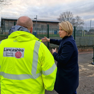 Katherine Fletcher on a school playground, talking to a man in a high-vis jacket. The jacket has the Lancashire County Council logo visible on the back.