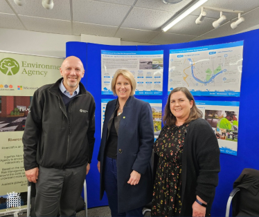 Katherine Fletcher stood with two members of the Environment Agency. An information board about the flood scheme is behind them.